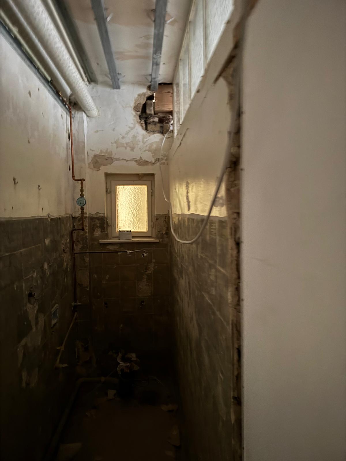 Schmaler Toilettenraum im Rohbauzustand mit alten Fliesen und Kupferrohren.